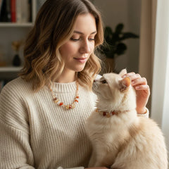 A woman and her cream-colored cat wearing matching orange and yellow faceted crystal necklaces, showing the warm bond between a pet and owner.